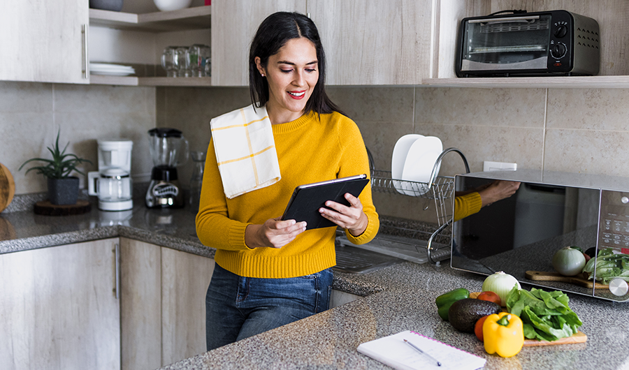 Mujer en la cocina, rodeada de electrodomésticos y mirando su tablet con interés 